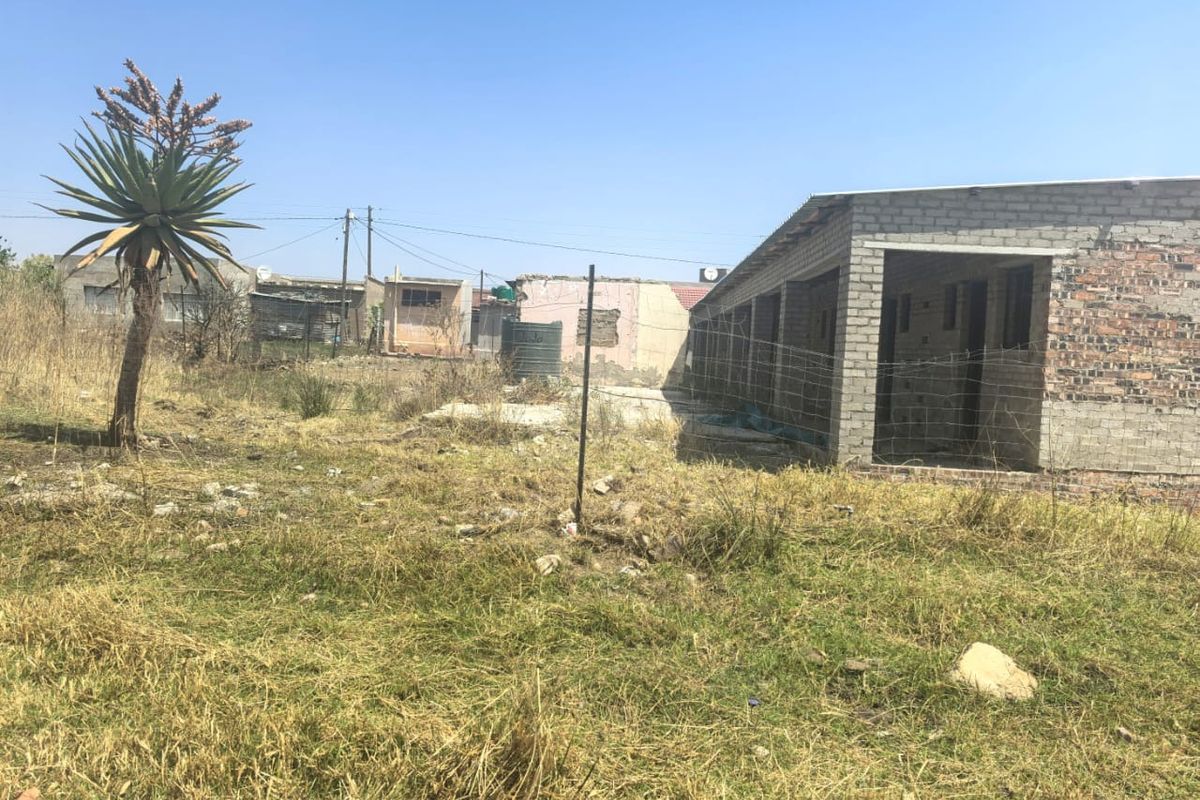 Rear view of an unfinished brick structure on a large plot with overgrown grass and a water storage tank.