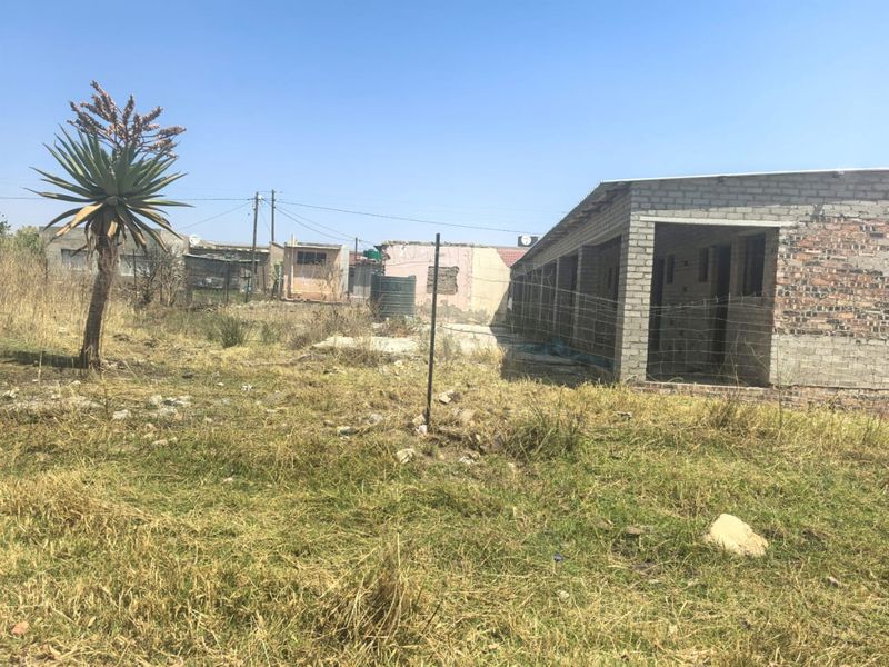 Rear view of an unfinished brick structure on a large plot with overgrown grass and a water storage tank.