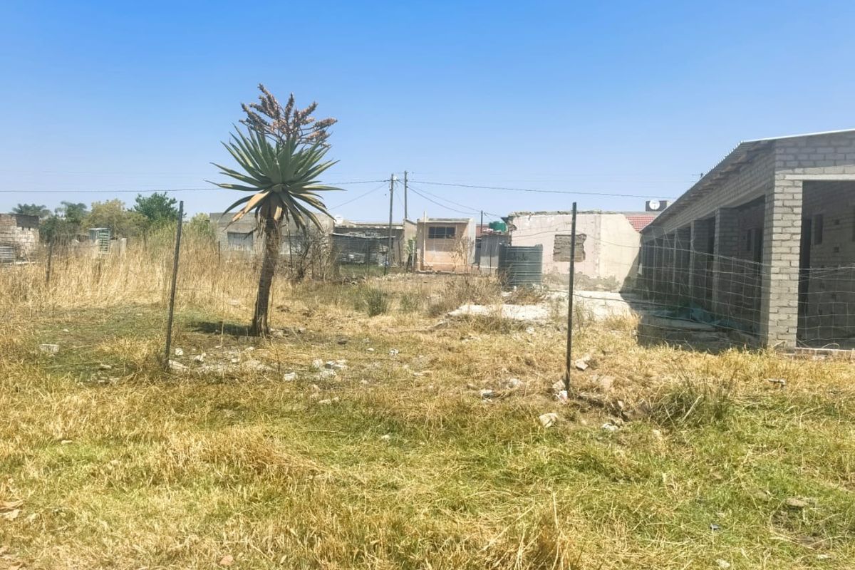 An exterior view of a property plot featuring a partially constructed concrete block structure, wire fencing, and a water storage tank on unmaintained land.