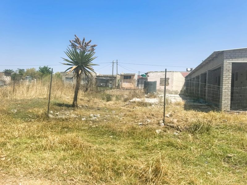 An exterior view of a property plot featuring a partially constructed concrete block structure, wire fencing, and a water storage tank on unmaintained land.