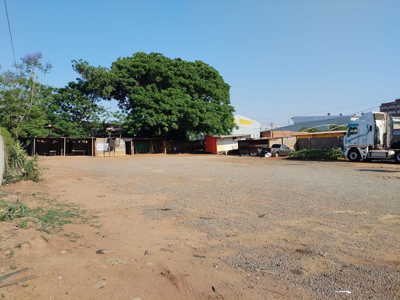 A large, open commercial vacant land plot featuring a gravel surface, perimeter walling, and mature trees with existing temporary structures on site.