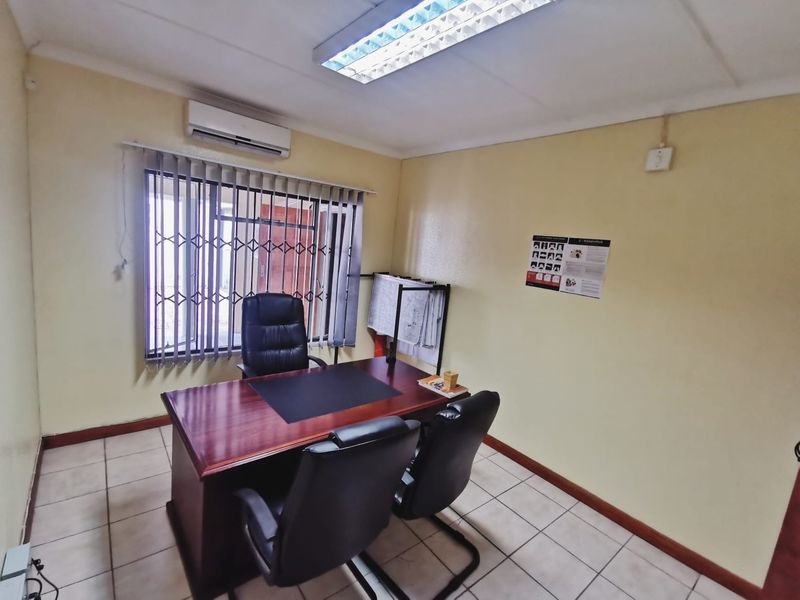 A functional office space featuring tiled flooring, a wall-mounted air conditioning unit, and a window fitted with security bars and vertical blinds.