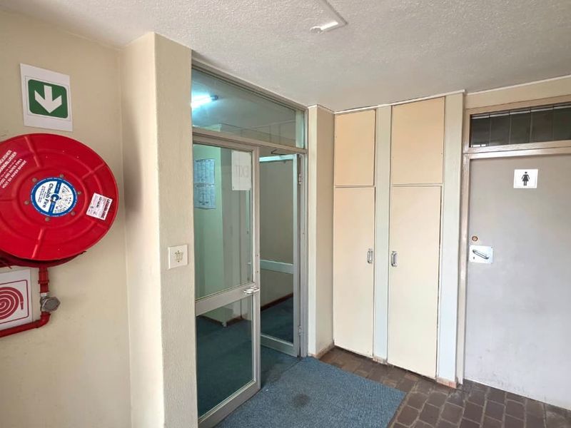 A functional office hallway featuring a wall-mounted fire hose reel, built-in storage cupboards, and access to a restroom facility.