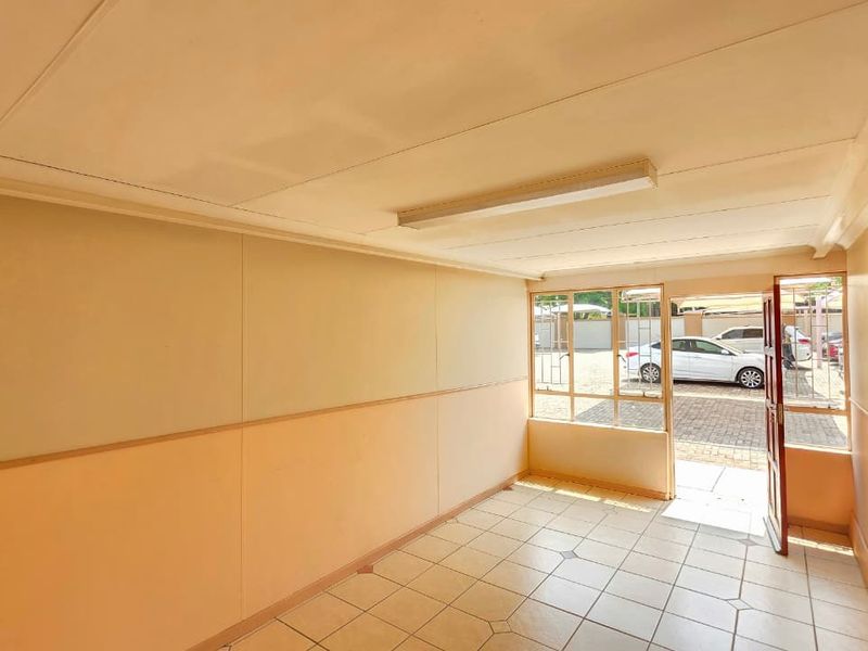 A bright, empty office space featuring tiled flooring, large windows providing natural light, and direct access to the exterior parking area.