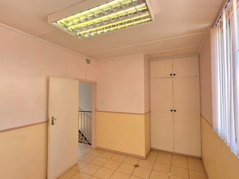 A functional office space featuring tiled flooring, built-in storage cupboards, vertical window blinds, and a recessed fluorescent light fixture.