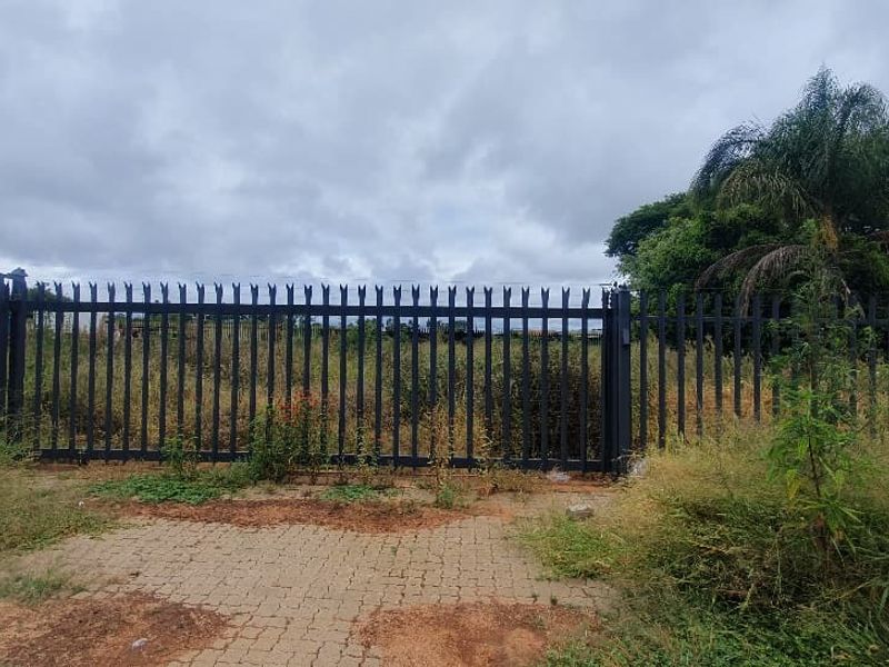 A view of a vacant commercial plot secured by metal palisade fencing and a gate, featuring a paved entrance area and overgrown vegetation.