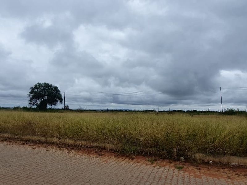 A view of a vacant commercial land plot featuring a paved road frontage, perimeter fencing, and open grassy terrain under a cloudy sky.