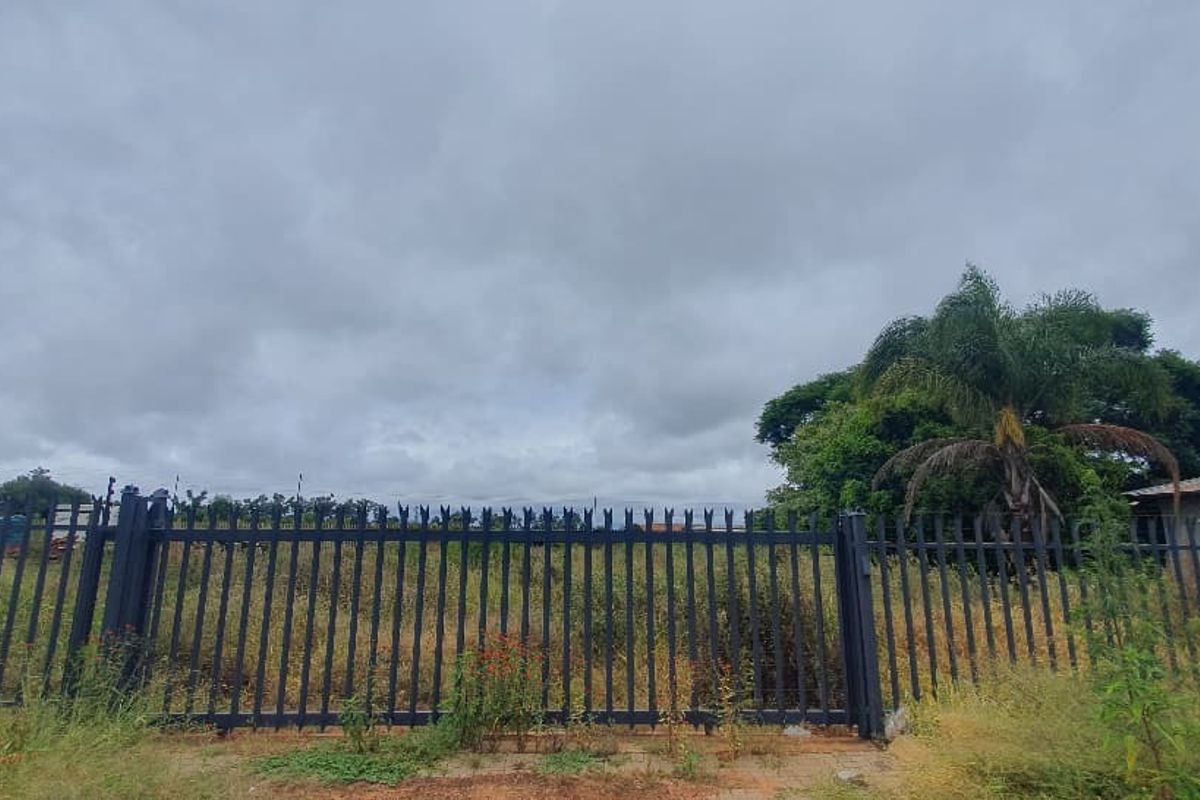 A view of a vacant commercial plot secured with metal palisade fencing and a gate, featuring overgrown vegetation on the site.