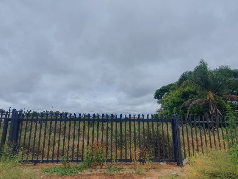 A view of a vacant commercial plot secured with metal palisade fencing and a gate, featuring overgrown vegetation on the site.