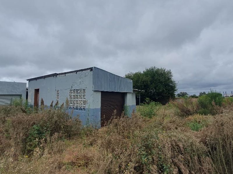 Exterior view of a commercial structure on a vacant lot, featuring metal roller doors and perimeter fencing, requiring significant maintenance and site clearing.