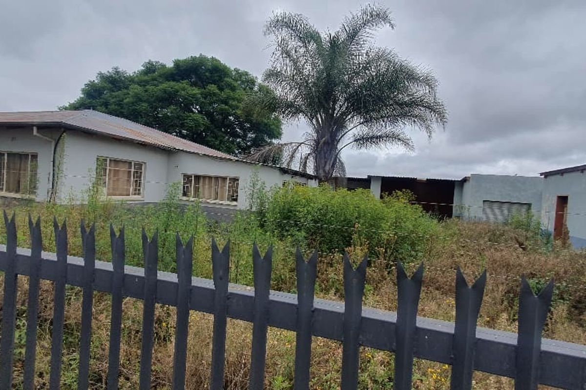 Street view of a commercial property featuring multiple structures, metal fencing, and significant overgrown vegetation requiring site clearance and maintenance.