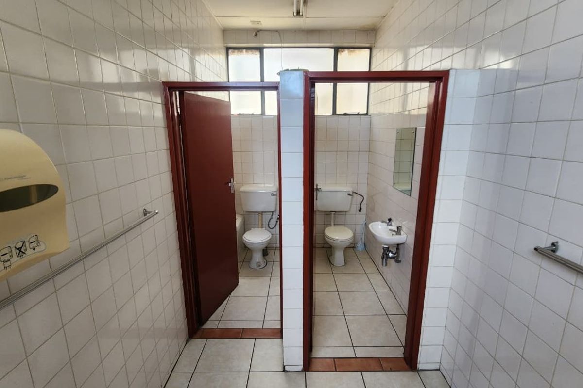 A functional office bathroom facility featuring two separate toilet cubicles, tiled walls and floors, and a small wall-mounted washbasin.