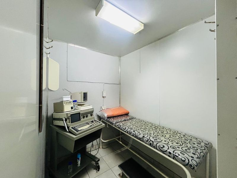A small clinical examination room featuring tiled flooring, wall-mounted hooks, and overhead fluorescent lighting, currently equipped with medical diagnostic equipment.