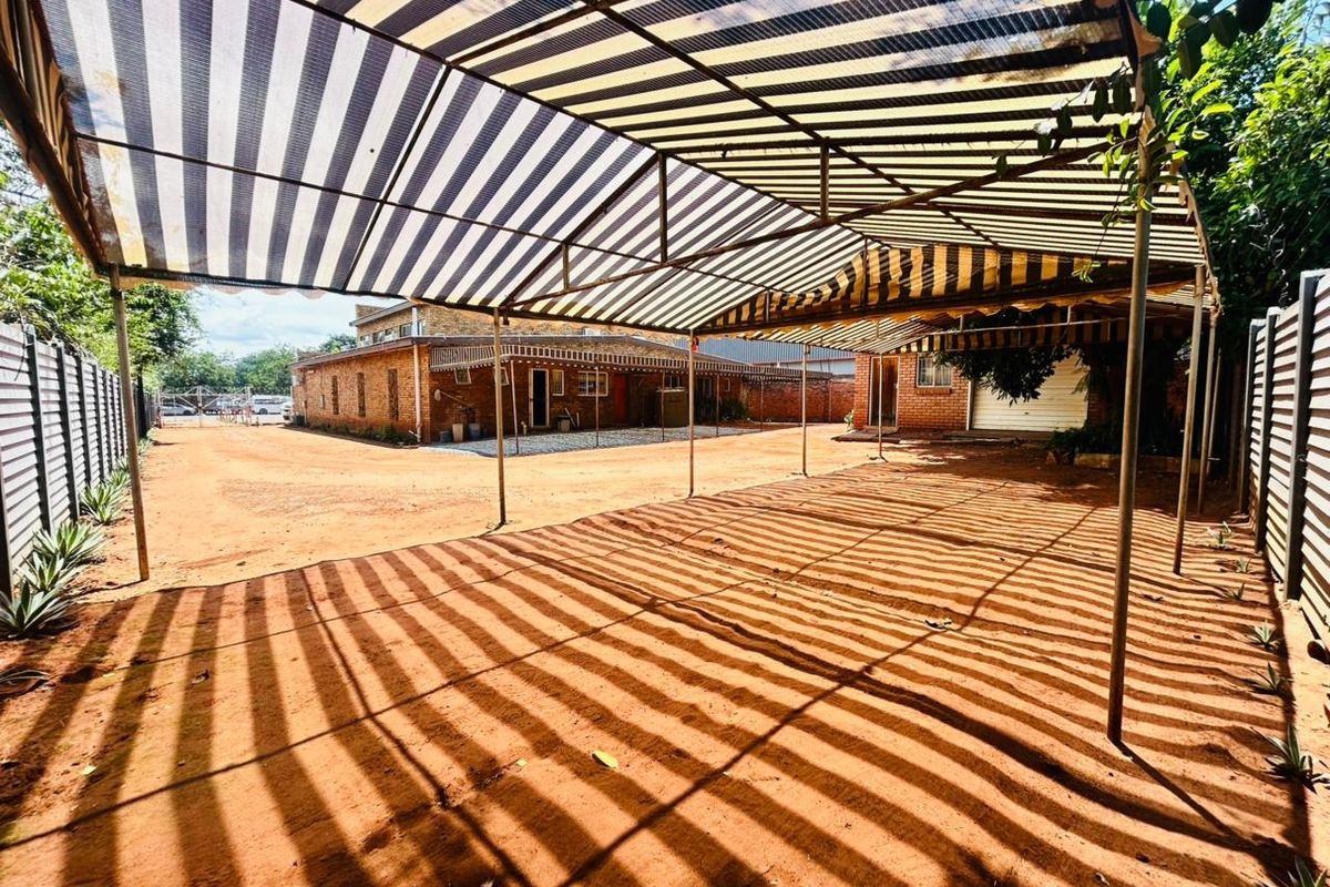 A large outdoor area featuring a metal-framed shade structure with striped cloth, overlooking a brick commercial building on an unpaved dirt lot.
