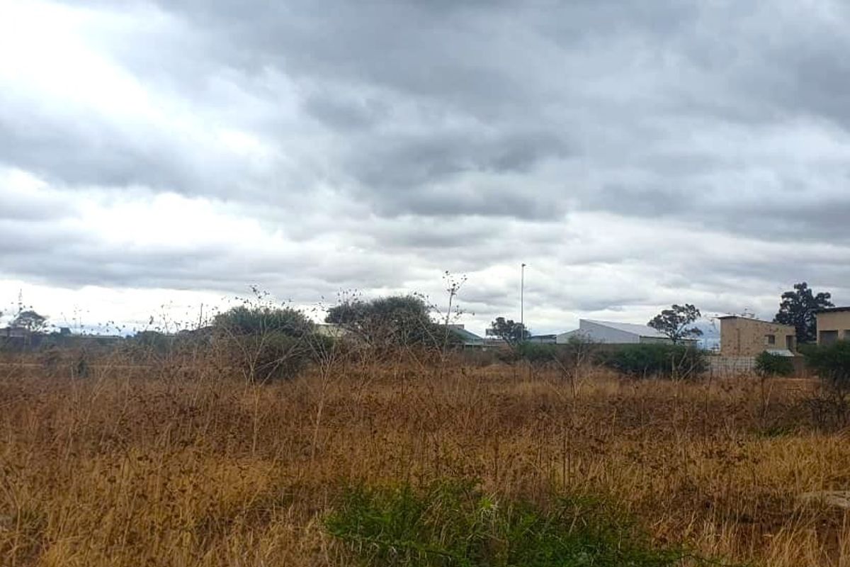 A view of a vacant commercial land plot featuring dry vegetation and surrounding neighborhood infrastructure under a cloudy sky.