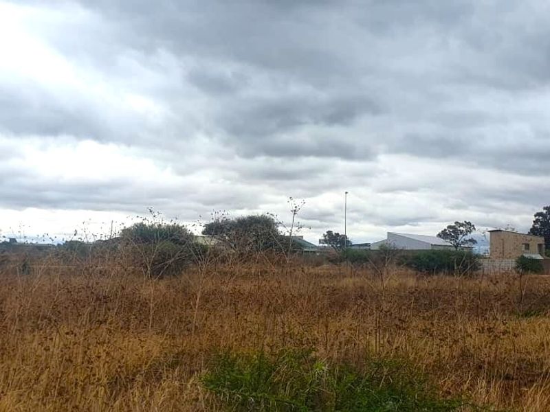 A view of a vacant commercial land plot featuring dry vegetation and surrounding neighborhood infrastructure under a cloudy sky.