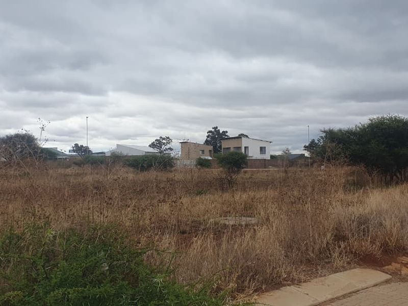 A view of a vacant commercial plot featuring overgrown dry grass and a paved verge, ready for development.