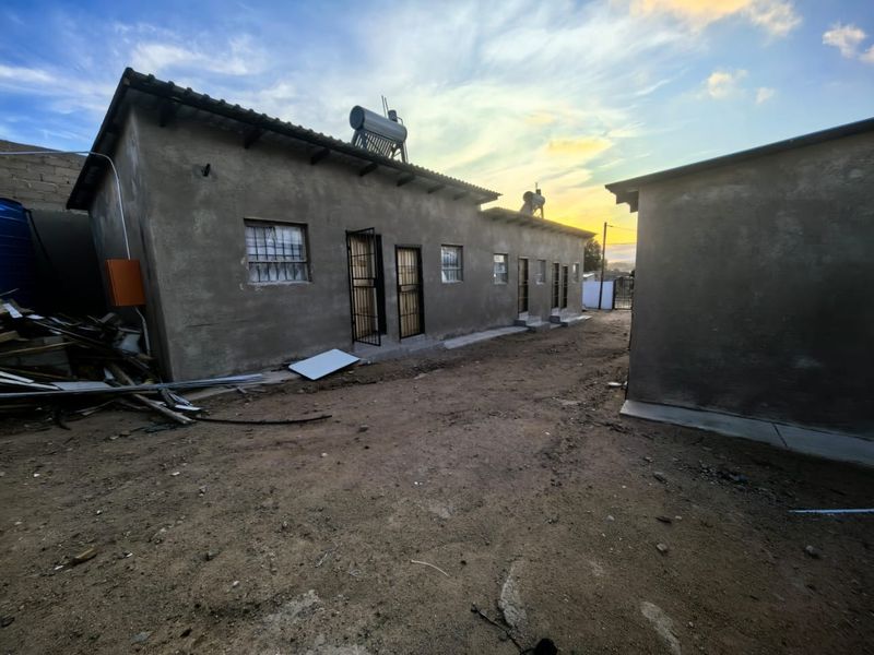 Rear exterior view of a multi-unit residential building featuring solar water heaters, security gates, and unfinished exterior wall surfaces with unpaved ground.