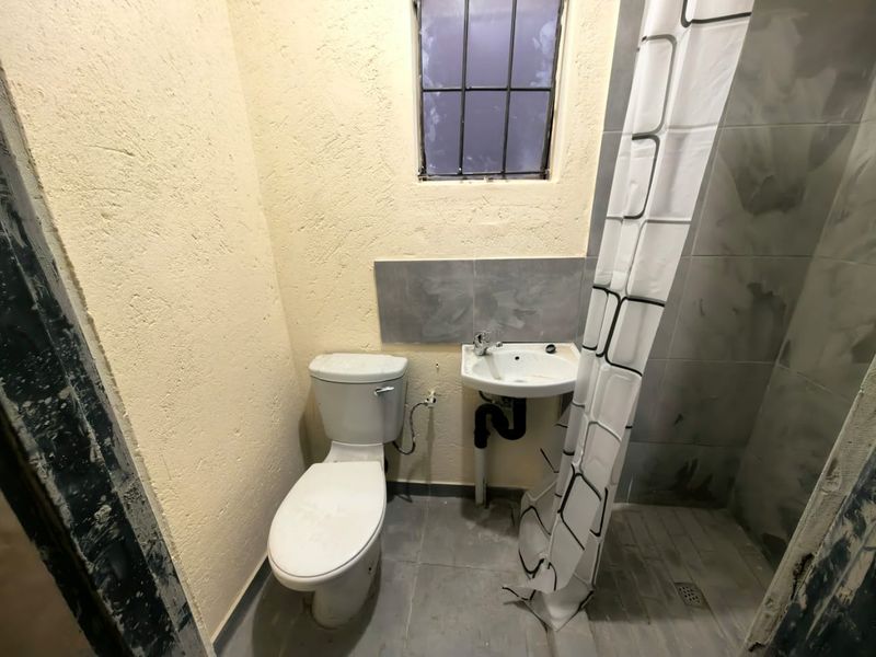 A compact bathroom featuring a toilet, wall-mounted basin, and a tiled shower area with exposed plumbing and unfinished wall surfaces.