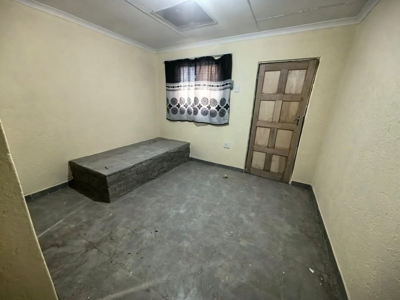 A basic, empty bedroom featuring tiled flooring, a built-in brick platform, and a wooden door, offering a blank canvas for renovation.