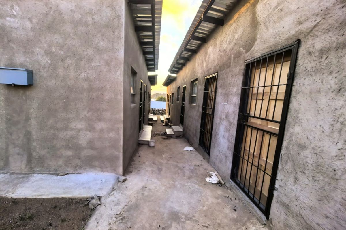 A narrow exterior walkway between two unpainted apartment structures featuring security gates and corrugated metal roofing.