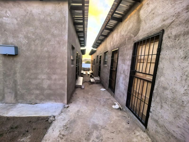 A narrow exterior walkway between two unpainted apartment structures featuring security gates and corrugated metal roofing.