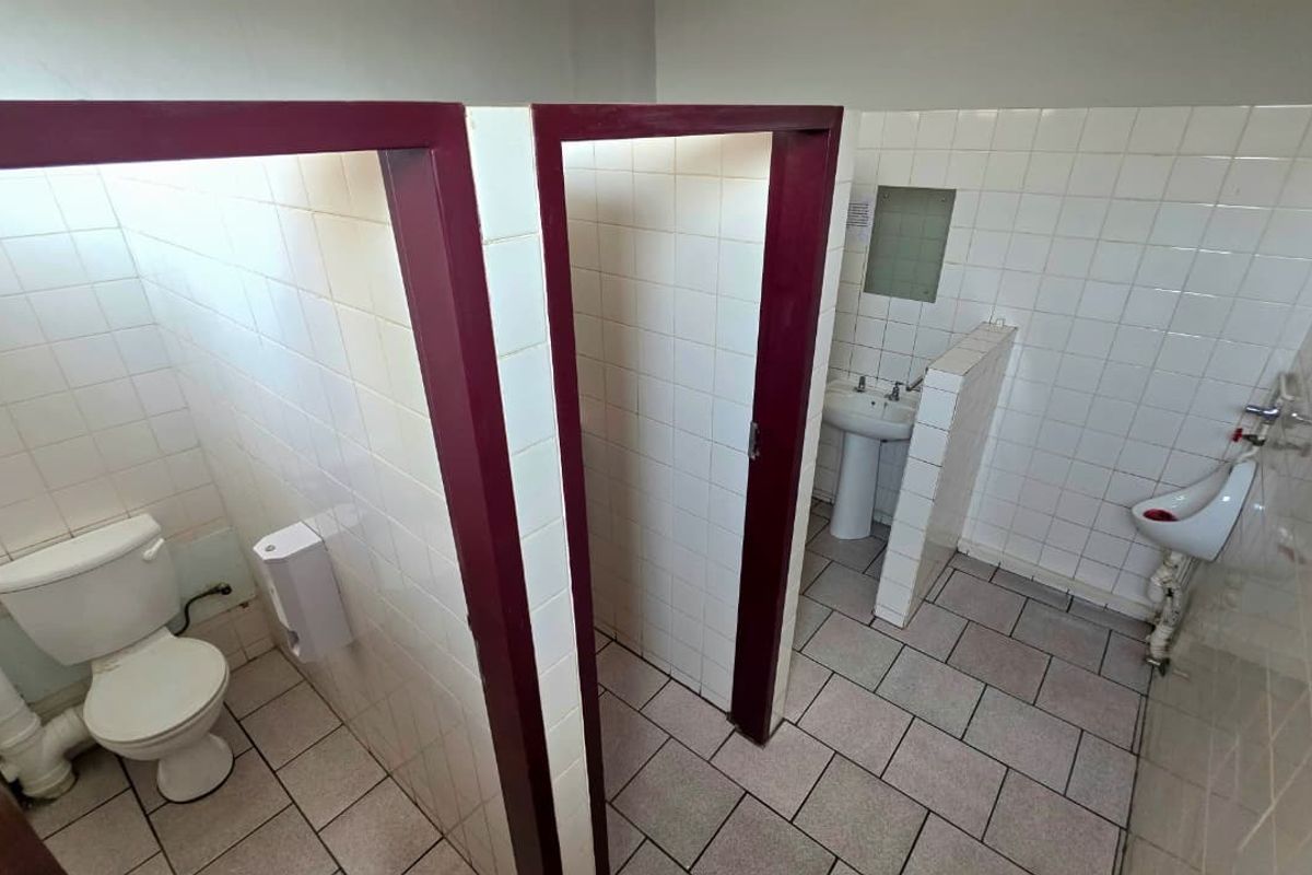A functional office bathroom featuring tiled walls and floors, equipped with a pedestal basin, urinal, and private toilet cubicles.