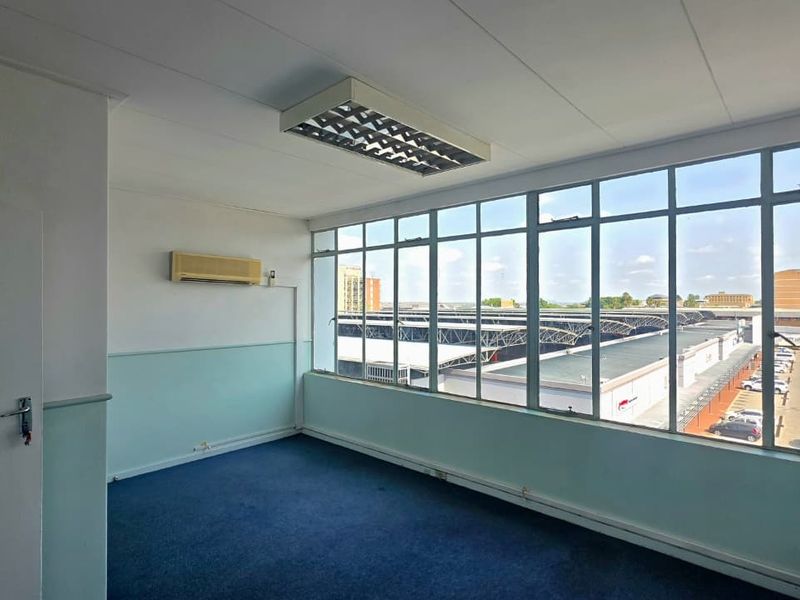 A vacant office space featuring large windows with industrial views, a wall-mounted air conditioning unit, and carpeted flooring.
