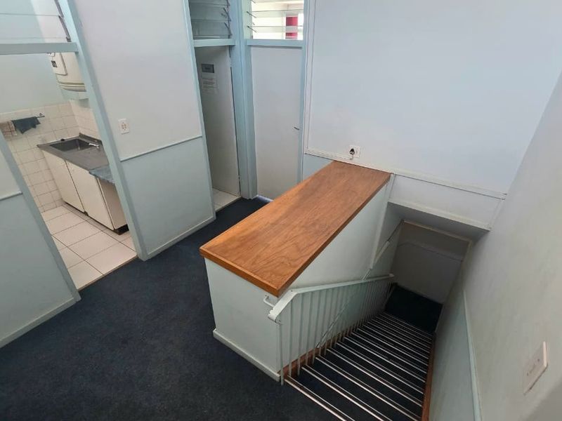 A functional office hallway featuring a staircase with a metal balustrade, adjacent kitchenette area, and louver windows for ventilation.
