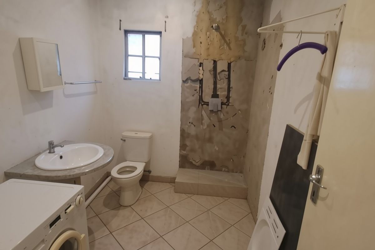 A bathroom in need of renovation, featuring a toilet, washbasin, and shower area with damaged wall finishes and exposed plumbing.
