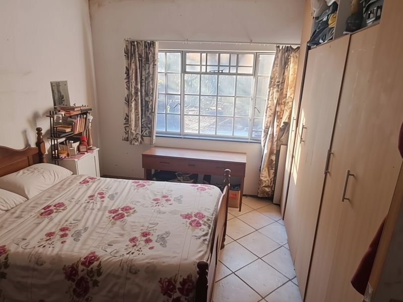 A bedroom featuring built-in cupboards, tiled flooring, and a window with security bars. The room requires cosmetic updates to the wall finishes.
