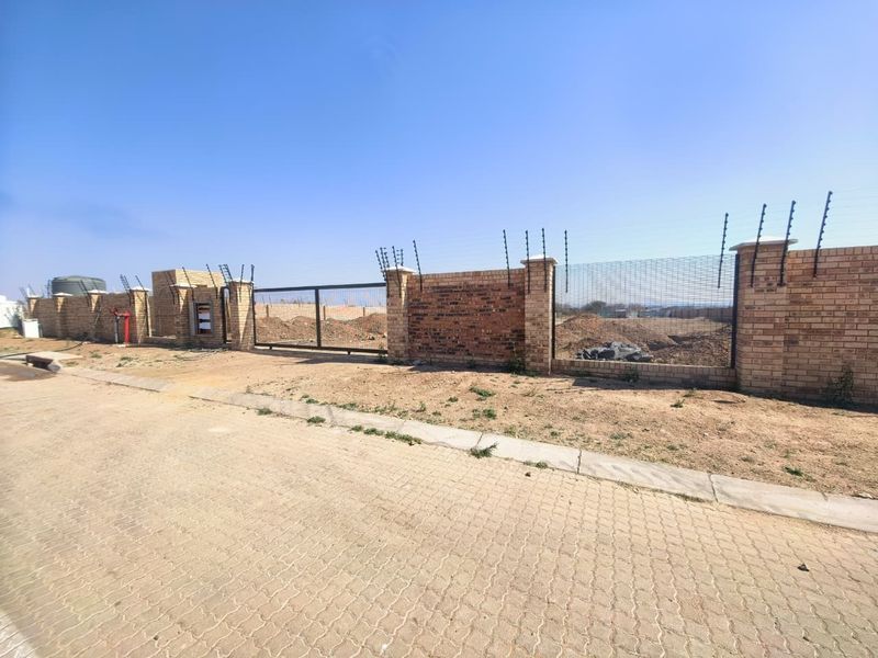 A vacant commercial land plot featuring a secure brick boundary wall, electric fencing, and a metal sliding gate, accessible via a paved street.