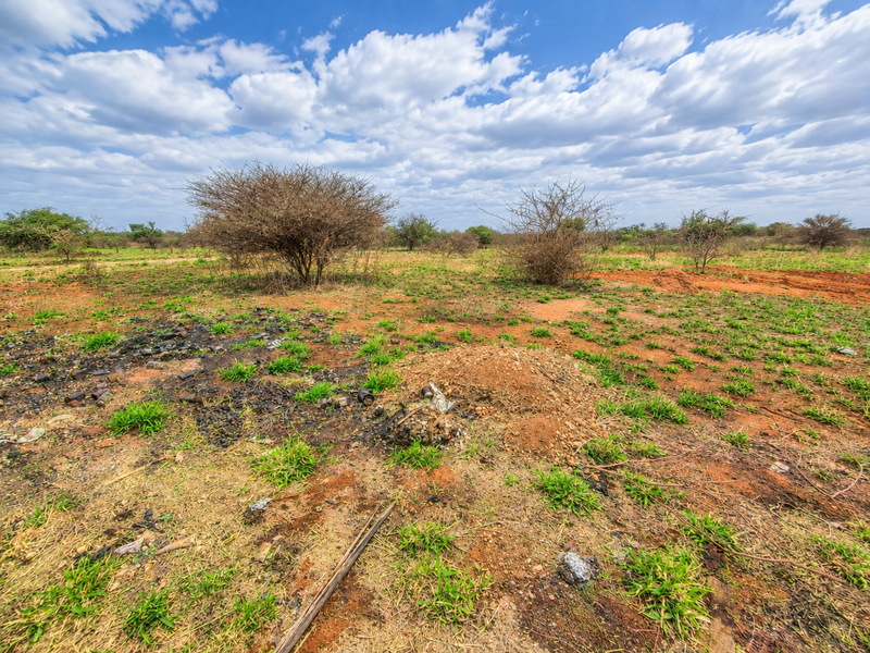 A view of a vacant residential plot featuring natural scrub vegetation and uneven earth terrain under a bright, cloudy sky.
