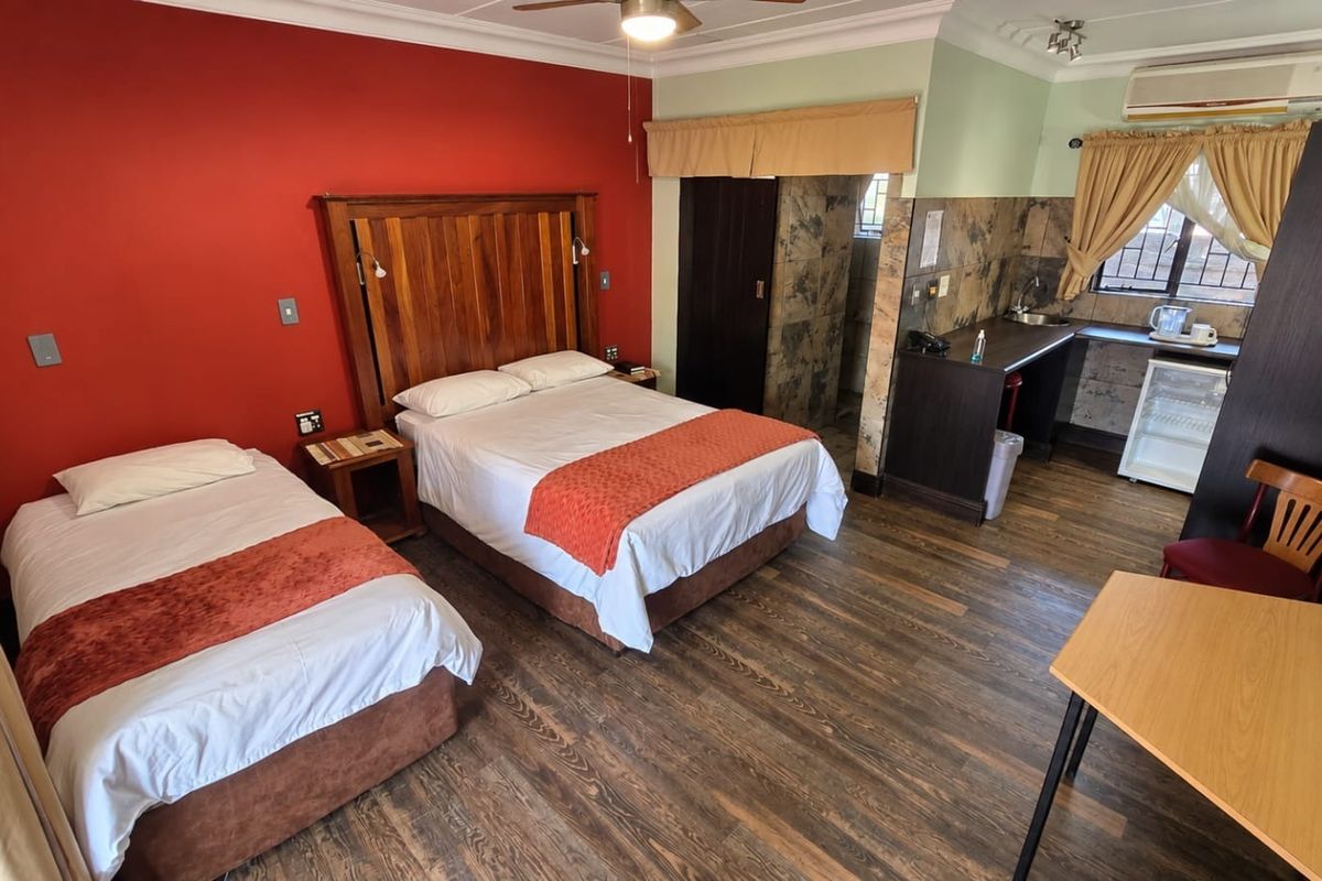 A hotel bedroom featuring a double and single bed, laminate flooring, and a compact kitchenette area with a sink and wall-mounted air conditioning.