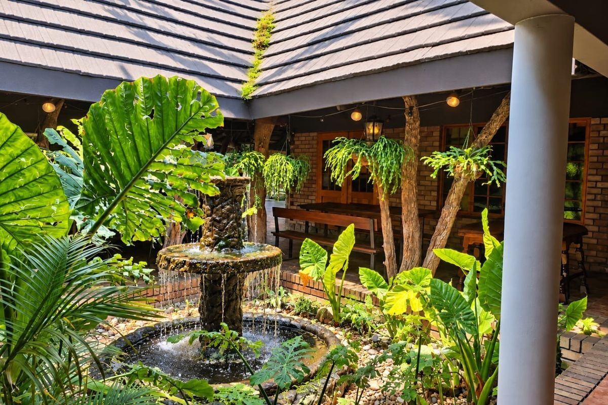 A tranquil covered patio area featuring a central stone fountain, brick wall finishes, and a tiled roof, surrounded by lush garden landscaping.
