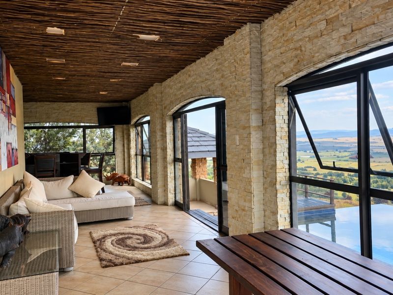 A spacious covered patio featuring stacked stone walls, a rustic thatched ceiling, and large aluminium windows offering expansive views over the landscape and pool.