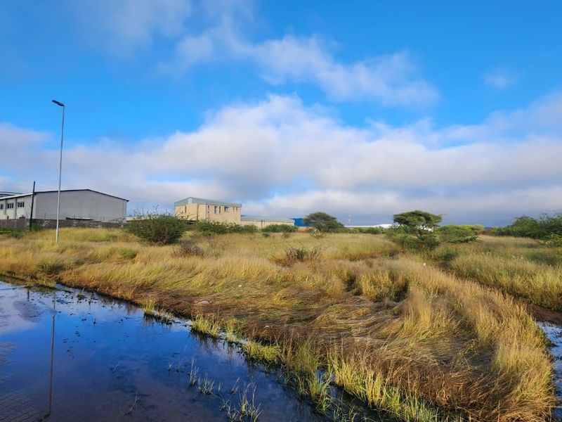 A view of a vacant commercial land plot featuring natural vegetation, paved road access, and nearby industrial structures under a clear sky.