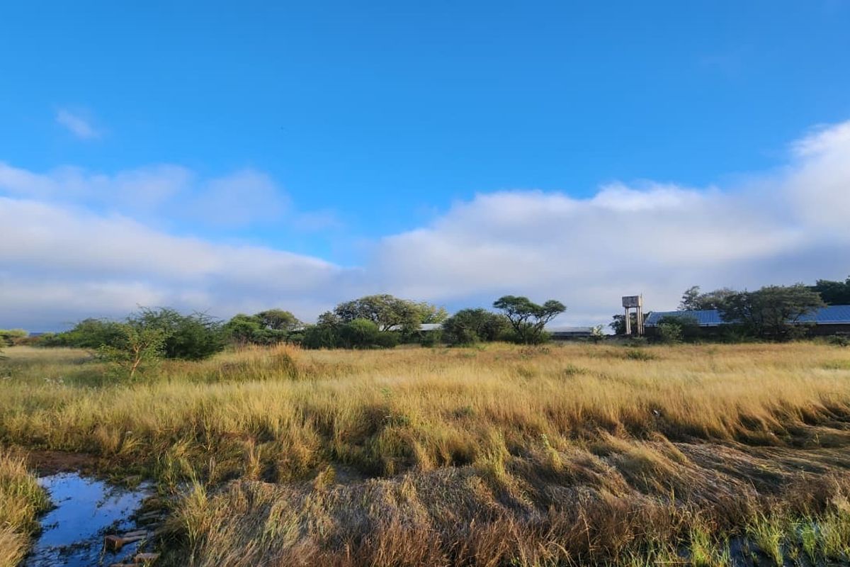 A wide view of the vacant commercial land featuring natural vegetation, a water tower, and existing metal roof structures in the distance.