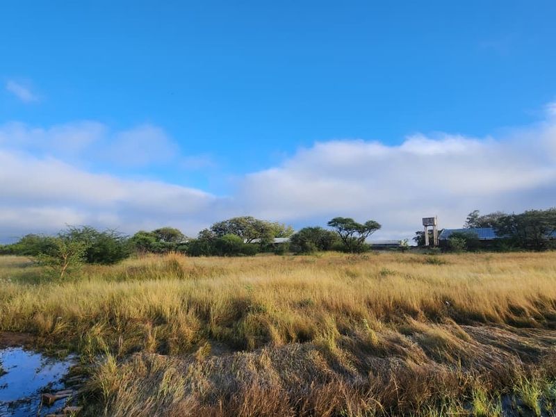 A wide view of the vacant commercial land featuring natural vegetation, a water tower, and existing metal roof structures in the distance.