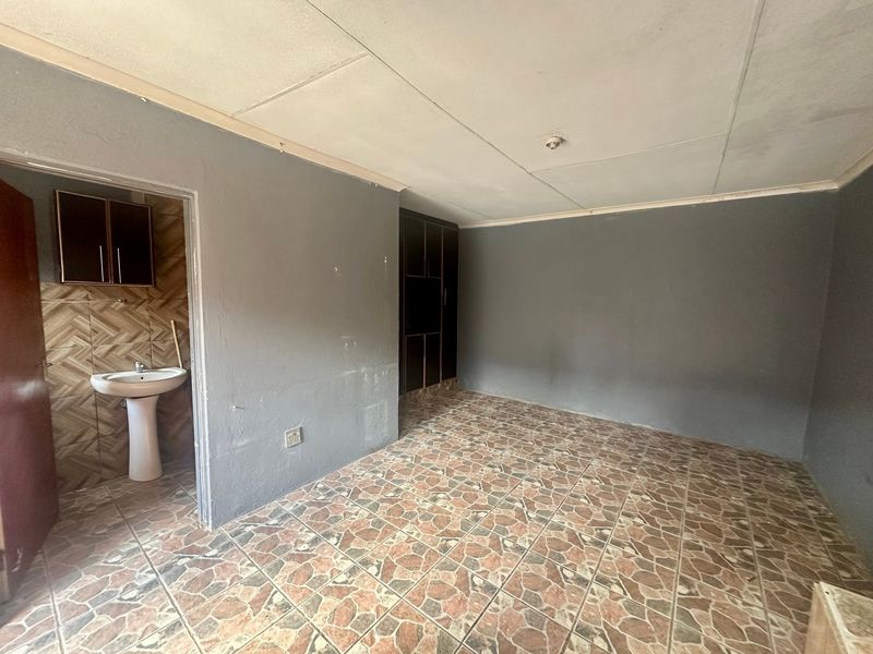 An empty bedroom featuring tiled flooring, built-in storage, and an attached en-suite area with a pedestal basin and wall-mounted cabinetry.