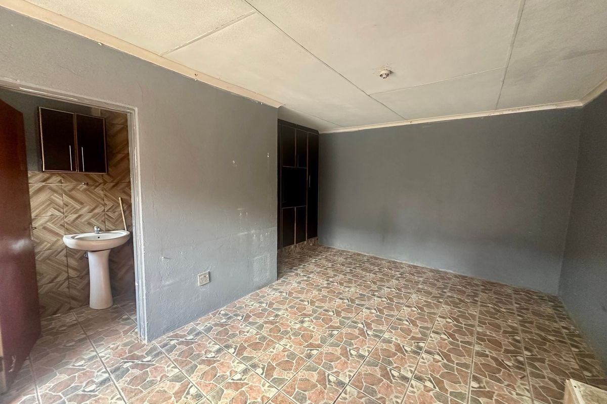 An empty bedroom featuring tiled flooring, built-in storage, and an attached en-suite area with a pedestal basin and wall-mounted cabinetry.
