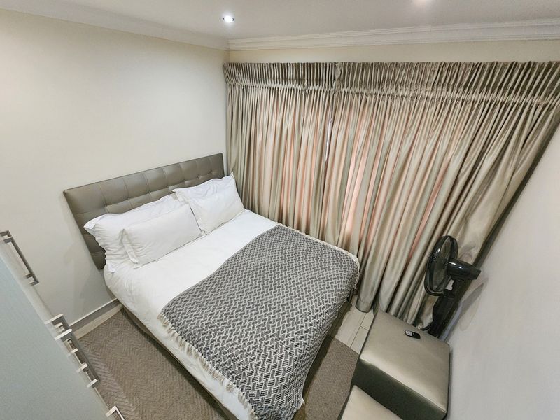 A compact, well-maintained bedroom featuring neutral wall tones, recessed ceiling lighting, and full-length window drapery.