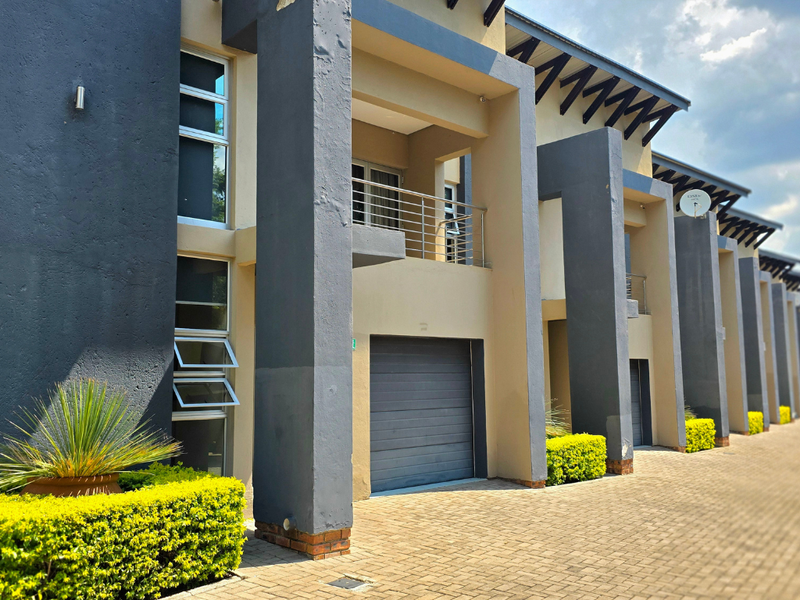 Modern townhouse exterior featuring a paved driveway, integrated garage, and upper-level balcony with contemporary architectural pillars and exposed rafter detailing.