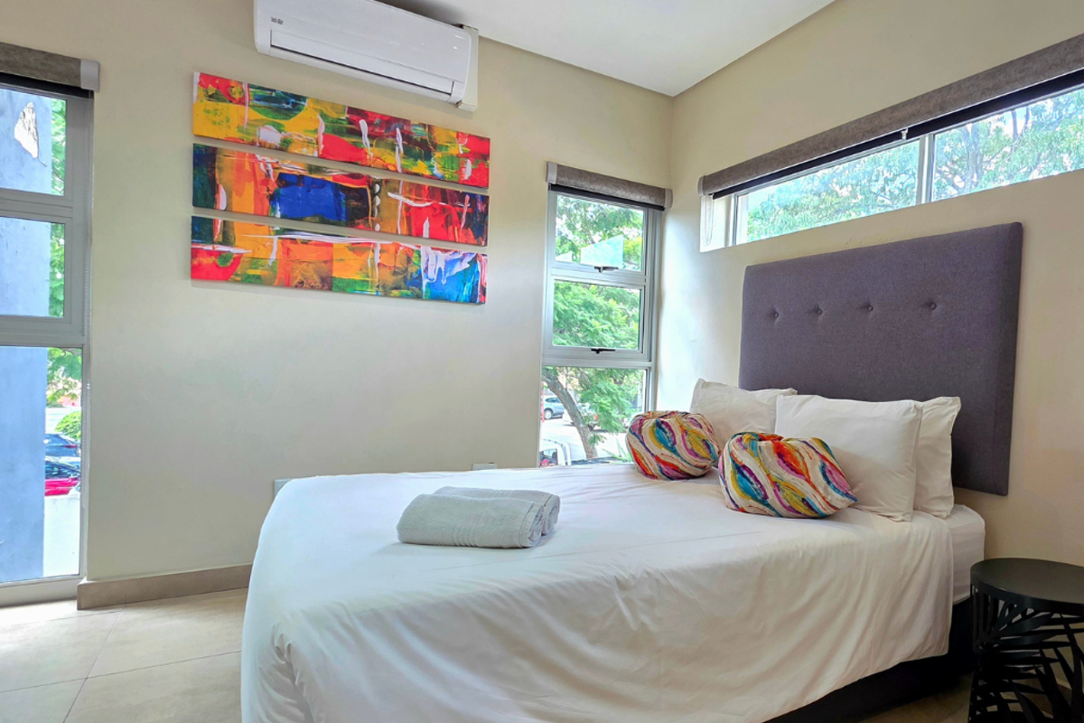 A well-lit bedroom featuring tiled flooring, a wall-mounted split-system air conditioner, and large aluminium-framed windows with roller blinds.