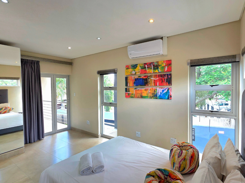 A well-maintained bedroom featuring tiled flooring, recessed lighting, a split-system air conditioner, and large aluminium-framed windows providing natural light and access to a balcony.
