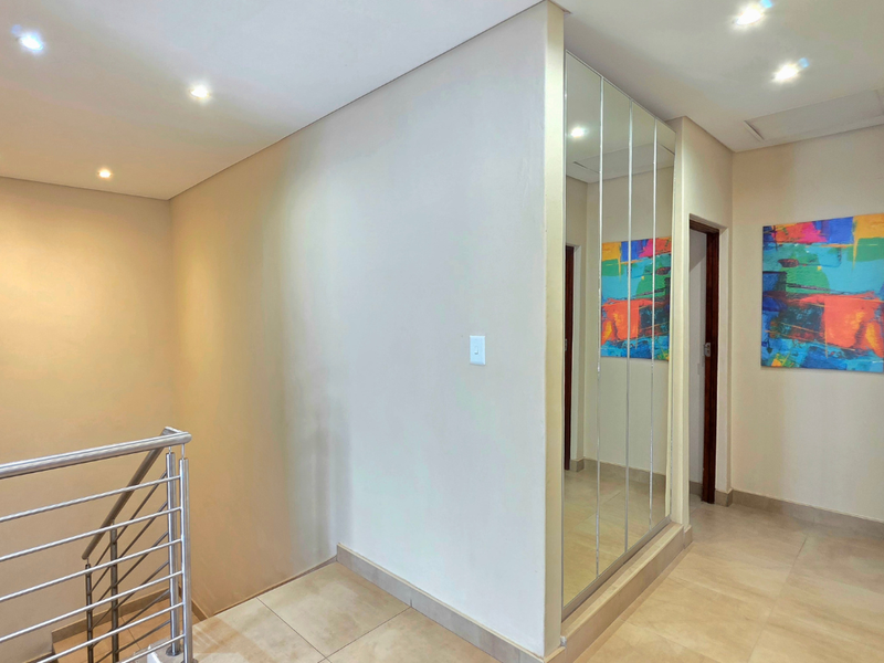A bright and modern hallway featuring tiled flooring, recessed downlighting, a stainless steel balustrade, and a large mirrored built-in cupboard.
