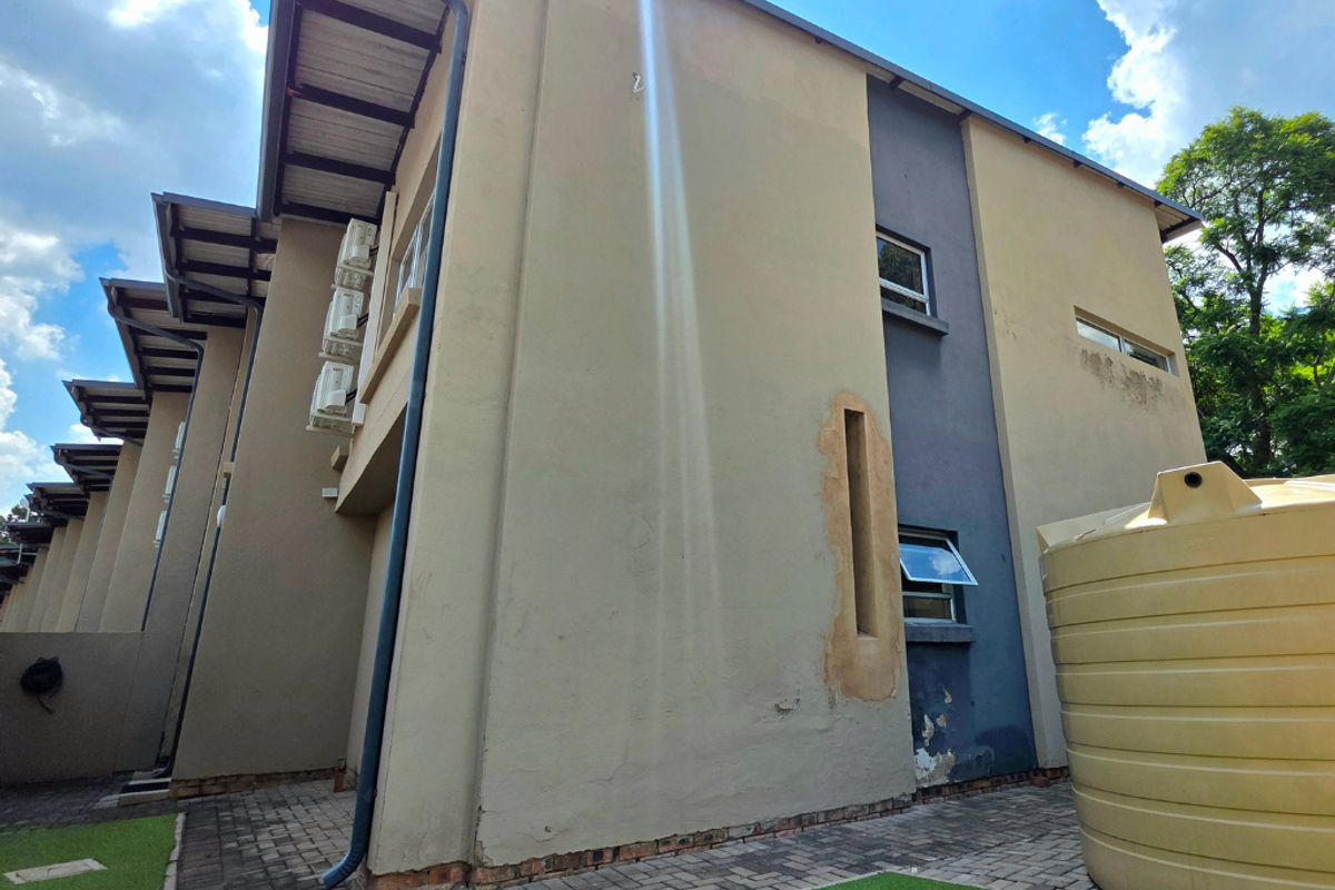 Rear exterior view of the townhouse featuring a water tank, paved pathways, and external air conditioning units with visible wear on the facade.