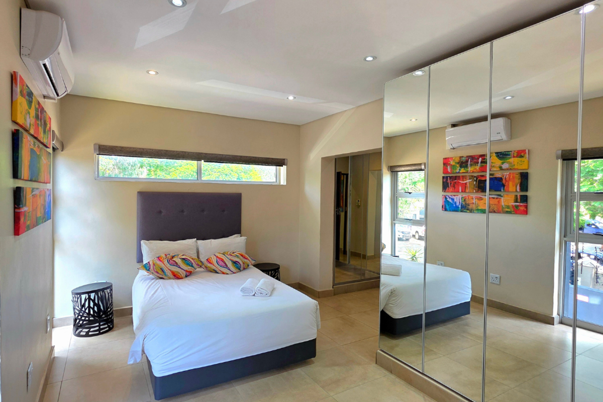 A modern bedroom featuring tiled flooring, recessed downlighting, a split-system air conditioner, and a large mirrored built-in wardrobe.