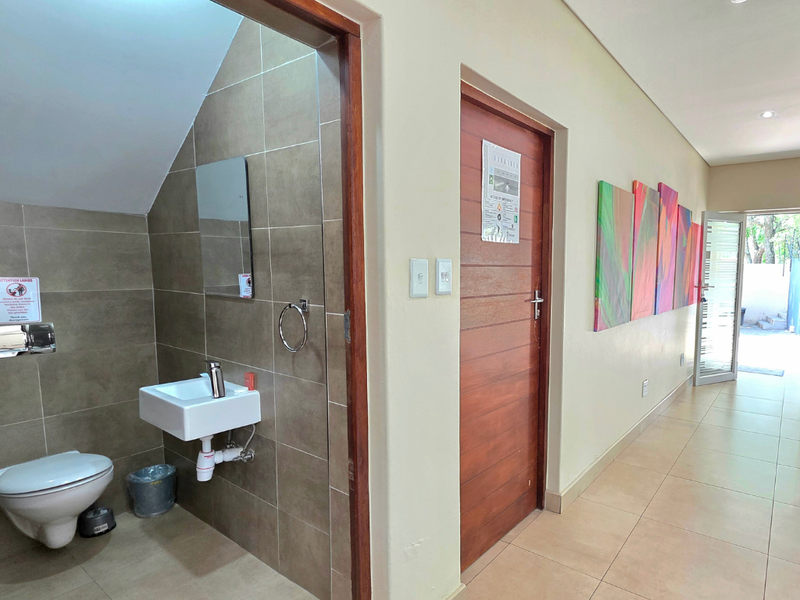 A clean, modern bathroom featuring a wall-mounted basin and toilet, complemented by neutral wall tiling and recessed lighting, adjacent to a tiled hallway.