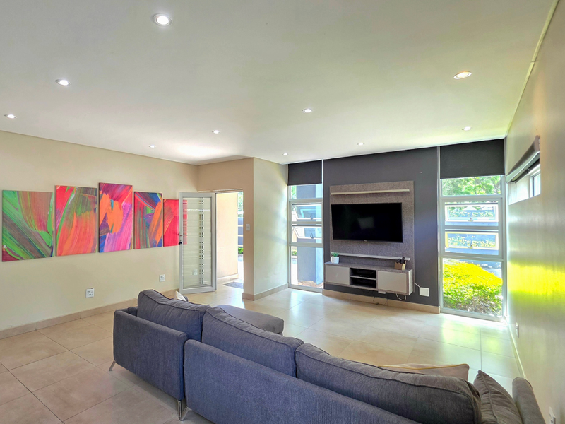 A bright and modern living room featuring tiled floors, recessed downlights, and a built-in media console with large windows overlooking the garden.